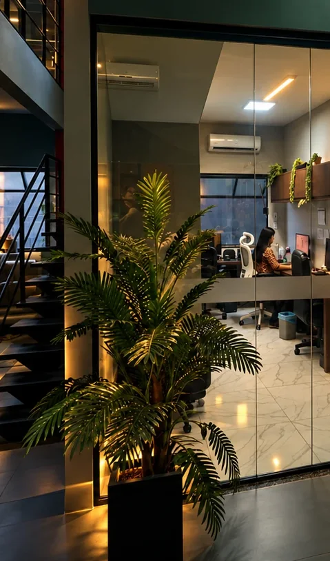 Atendente trabalhando em um escritório com porta de vidro, planta ornamental e escada iluminada, à noite